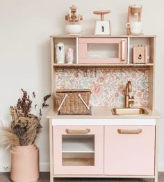 a pink wooden play kitchen with lots of storage and decor on the top shelf, next to a potted plant