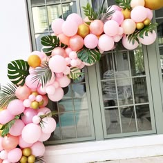 a pink and gold balloon arch in front of a building with palm leaves on it