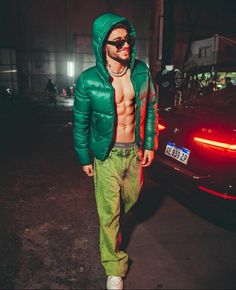 a man with no shirt and green pants standing in front of a car at night