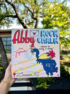 a hand holding up a rock chalk book in front of a tree and building with the words rock chalk written on it