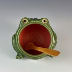 a green ceramic frog cup with a wooden spoon in it's mouth and eyes