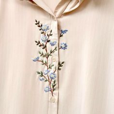 a white shirt with blue flowers embroidered on the chest and collar, hanging from a wooden hanger