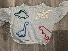 a white sweater with colorful dinosaurs on the front and back, sitting on top of a wooden floor