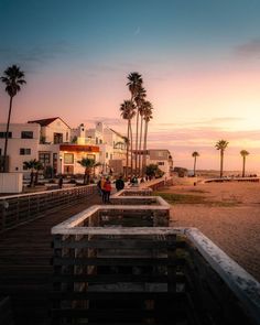 people are walking on the boardwalk near some buildings and palm trees at sunset or dawn