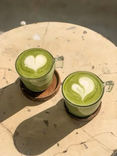 two cups of green tea with white hearts in them sitting on a wooden table next to each other