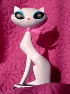 a white cat figurine sitting on top of a pink bed sheet with blue eyes