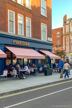 people are sitting at tables in front of a restaurant on the side of the street