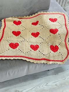 a crocheted afghan with red hearts on it sitting on a gray couch next to a pillow