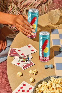 a table topped with two cans of beer and playing cards next to a bowl of popcorn