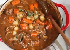 a pot filled with stew and carrots on top of a white table cloth next to a wooden spoon