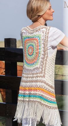 a woman wearing a crocheted vest and white jeans standing on a wooden fence