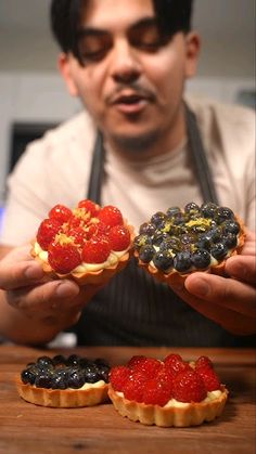 a man holding three pastries in front of him on a wooden table with fruit