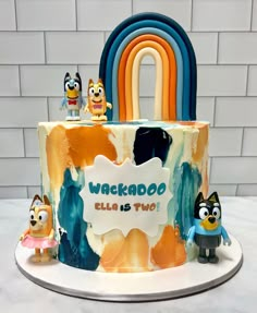 a birthday cake decorated with cartoon characters and a rainbow shaped sign that says wakabo ella is two