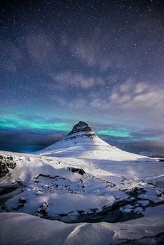 the sky is filled with green and blue lights as it stands on top of a snow covered mountain