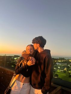 two people standing next to each other on top of a roof at sunset with the city lights in the background