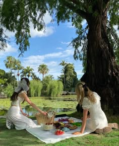 two women in white dresses are sitting on a blanket near a tree and picnicking