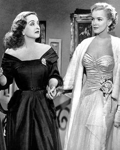 two women dressed in evening gowns standing next to each other, one wearing a cape