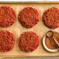 four hamburger patties on a baking sheet with seasoning in a bowl and spoon