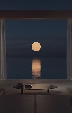 a full moon is seen over the ocean from a living room with white drapes