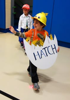 two children wearing hard hats and holding a hatch sign