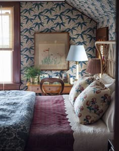 a bed sitting under a window next to a lamp and table in a room with blue wallpaper