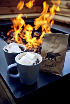 two cups of hot chocolate sitting on top of a table next to an open fire pit