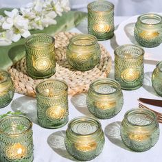 many green glass candles are sitting on a white table cloth with flowers in the background