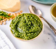 a bowl filled with green pesto next to two spoons on a white table