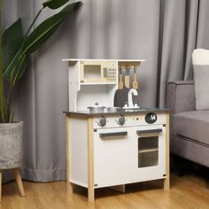 a white wooden play kitchen set in a living room with a gray couch and potted plant