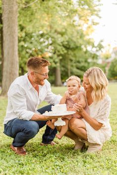 a man and woman holding a baby in front of a cake while sitting on the grass