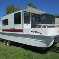 a white boat sitting in the grass next to a trailer with a house on it