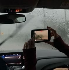 a person holding up a cell phone in front of a car with raindrops on the windshield