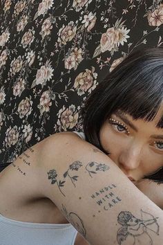 a woman with tattoos on her arm sitting in front of a flowered wallpaper