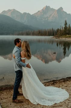 a bride and groom standing on the shore of a mountain lake