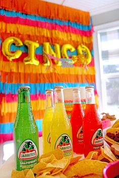 three bottles of soda, chips and salsa on a table in front of a party banner