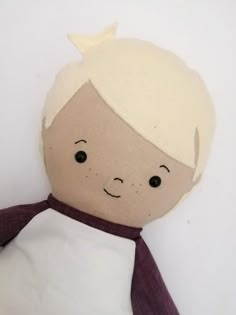 a stuffed doll with a white shirt and purple scarf on it's head, sitting against a white wall