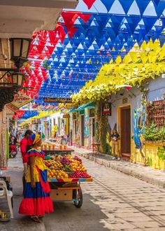 a woman standing next to a cart filled with fruit under colorful umbrellas on the side of a street