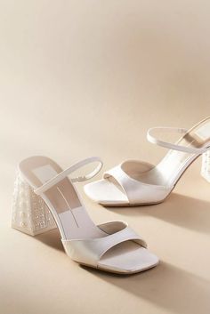 two pairs of white high heeled shoes on top of a beige surface, one in the foreground and the other in the background