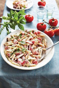 a white bowl filled with pasta salad next to tomatoes and green leaves on the table