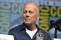 a bald man in a blue shirt is at a microphone and looks off into the distance