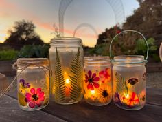 four mason jars filled with flowers and lit candles on a wooden table in front of a sunset