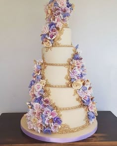 a three tiered wedding cake with purple flowers on the side and gold trimmings