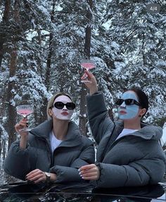 two women with face paint holding up wine glasses in the snow while sitting on top of a car