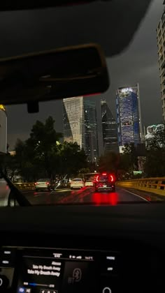 a car driving down a street with tall buildings in the background at night, seen from inside a vehicle's dashboard