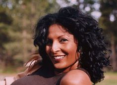 a woman with black curly hair smiling at the camera while sitting on a bench in a park