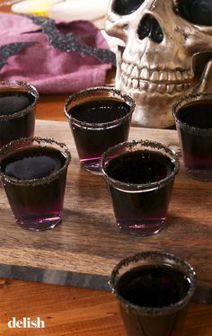 six shot glasses filled with dark liquid sit on a wooden table next to a skull