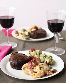 two white plates topped with meat and shrimp next to wine glasses filled with red wine