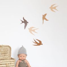 a small doll sitting next to a wicker chair and wall with birds on it