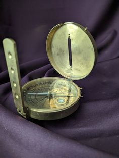 an open brass compass sitting on top of a purple cloth next to a metal object