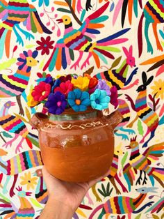 a hand holding a clay pot with flowers in it on a colorful wallpaper background